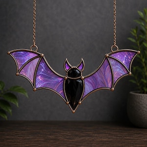 May include: A hanging bat ornament featuring a central black faceted body and ears, with wings crafted from purple and violet swirled glass. The piece is framed in a dark metallic border and suspended by two thin chains attached to the wing tips.
