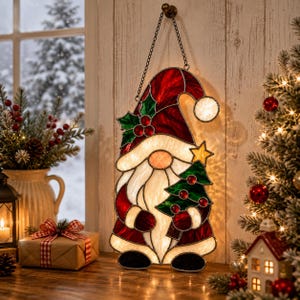 May include: A stained glass hanging ornament shaped like a gnome wearing a red hat with green holly leaves and red berries. The gnome has a white beard, holds a green Christmas tree with red ornaments and a yellow star, and hangs from a metal chain.