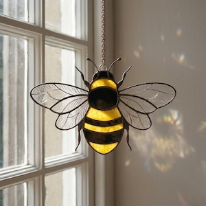 May include: A stained glass bee suncatcher with yellow and black stripes on the body and clear wings. The bee is suspended by a chain, perfect for hanging in a window. The suncatcher is a decorative item.