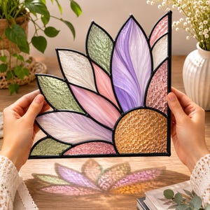 May include: A stained glass suncatcher shaped like a flower quarter-circle, featuring textured glass petals in shades of lavender, pink, white, and green, with a central textured amber glass piece, all held together by black metal solder lines.