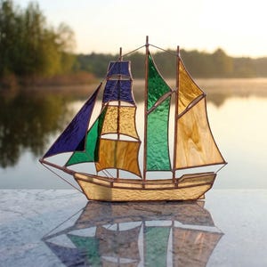 Könnte beinhalten: Ein Segelboot aus Buntglas mit cremefarbenem Rumpf und bunten Segeln. Die Segel bestehen aus blauem, grünem und gelbem Glas mit einem kupferfarbenen Rahmen. Das Segelboot spiegelt sich in der darunter liegenden Oberfläche.