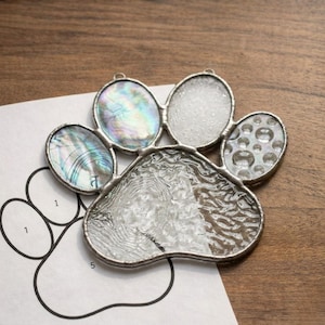 May include: A stained glass paw print with iridescent and textured glass pieces. The paw print is made of clear glass with a silver solder frame. The paw print is on top of a paper pattern with the numbers 1 and 5.