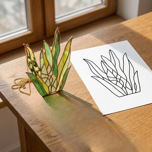 May include: A stained glass succulent pendant with a gold chain. The pendant features shades of green and yellow glass, with a detailed design. A black and white line drawing of the succulent is next to the pendant on a wooden surface.