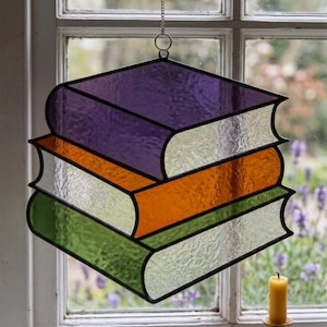 May include: A stained glass suncatcher shaped like a stack of three books. The top book is purple, the middle is orange, and the bottom is green. Each book features textured clear glass on the side panels, all held together by black metal cames.