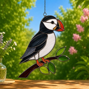 May include: A stained glass suncatcher depicting a puffin with black and white plumage, an orange and black beak, and orange feet, perched on a brown branch with green leaves. The piece is suspended by a metal chain against a blurred outdoor background.