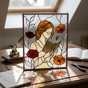 May include: Stained glass panel featuring a woman with red hair reading a book, surrounded by red and orange poppies and foliage. The artwork is framed in a dark brown border and is designed to hang.