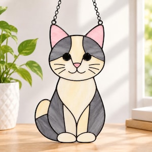 May include: A stained glass suncatcher shaped like a sitting cat with grey, cream, and pink glass panels. The cat features black glass eyes, a pink nose, and black wire whiskers, suspended by a black metal chain for hanging in a window.