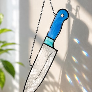 May include: A stained glass suncatcher shaped like a chef's knife, featuring a textured clear glass blade and a bright blue handle with a small turquoise accent. The piece is framed in black metal and hangs from a black chain with a suction cup.
