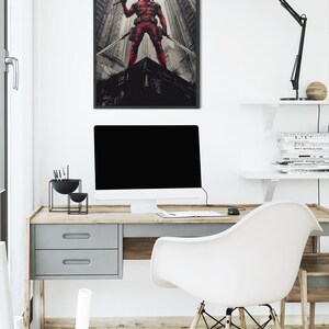 May include: A framed print depicting a red costumed figure with two swords, posed on a rooftop, with a cityscape backdrop. The print hangs above a desk with a computer and a white chair. The buildings are in shades of grey.