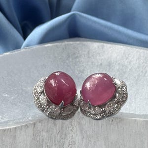 May include: A pair of stud earrings featuring large, round, deep pink gemstones. Each gemstone is surrounded by a halo of smaller, clear stones set in a silver-colored metal. The earrings are displayed on a light-colored surface.