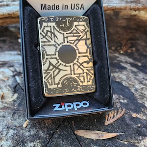 May include: A gold-toned Zippo lighter with a geometric design and a black center circle, presented in a black box. The lighter has a complex pattern and the text "Made in USA" is visible.