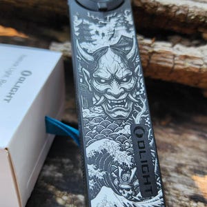 May include: A black rectangular Olight torch featuring a detailed white graphic of a horned demon mask, stylised waves, and a warrior figure. The device has a circular blue metallic button at the top and the brand name Olight printed vertically.