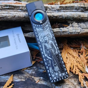 May include: A black Olight ArkPro flashlight featuring a detailed engraved graphic of a character holding a knife, surrounded by skull motifs. The flashlight has a circular blue button and is positioned next to its white product box.