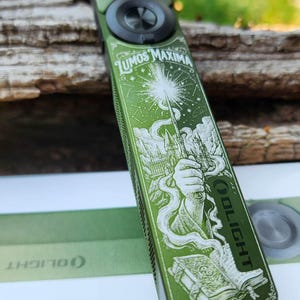 May include: A rectangular green Olight ArkPro flashlight featuring an engraved illustration of a hand holding a wand casting a spell with the text Lumos Maxima, a castle, and an open book. The flashlight has a black circular button at the top.