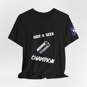 May include: A black short-sleeve t-shirt featuring a graphic of a 10 mm socket tool in the center. The text HIDE & SEEK is printed above the socket and the word CHAMPION is printed below it in white. A small blue logo is on the right sleeve.