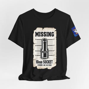 May include: A black short-sleeve t-shirt featuring a graphic of a missing 10mm socket on a beige paper background. The design includes a measurement scale, the text MISSING 10mm SOCKET, and REWARD: ONE SIX-PACK. A small blue logo is on the sleeve.