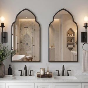 Op de afbeelding: Twee gewelfde wandspiegels met zwarte frames boven een dubbele wastafel. De spiegels hebben een spits toelopend ontwerp en reflecteren een badkamerinterieur met marmeren tegels, een badkuip en ingebouwde planken.