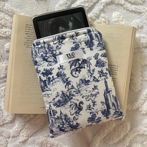 May include: A white and blue dragon-themed e-reader case with the text "TLC Threads". The case is open, revealing an e-reader. The case is placed on top of an open book with white pages.