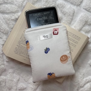 Könnte beinhalten: Weiße E-Book-Hülle mit einem Muster aus Pfannkuchen, Blaubeeren und einer Kaffeetasse. Die Hülle hat ein kleines Etikett mit der Aufschrift "TLC Threads". Ein E-Reader zeigt das Buch "Empire of Storms" von Sarah J. Maas an.