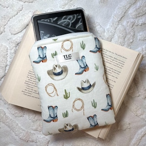 May include: A white ebook reader case with a zip closure, patterned with cowboy boots, hats, cacti, and rope designs. The case has a tag that reads "TLC Threads". An ebook reader is inside the case, resting on an open book.
