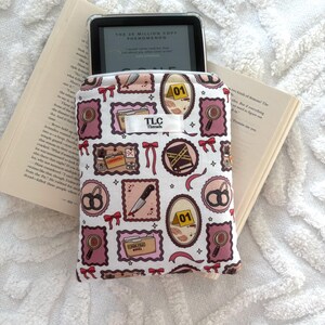 Può includere: Una custodia Kindle a fantasia bianca e rosa a tema detective, con lenti d'ingrandimento, coltelli e la scritta "TLC Threads". La custodia è aperta, rivelando un e-reader che mostra il testo di un libro.