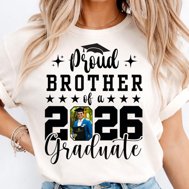 Puede incluir: Camiseta color crema con el texto "Proud Brother of a 2026 Graduate". El dise&ntilde;o incluye un gorro de graduaci&oacute;n, estrellas y una foto de un graduado con una blazer azul sosteniendo un diploma. Un regalo para un hermano orgulloso.