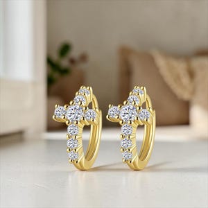 May include: A pair of gold-colored hoop earrings, each adorned with a cross design. The crosses are embellished with multiple clear, round-cut gemstones. The earrings are displayed on a white surface, with a blurred background.