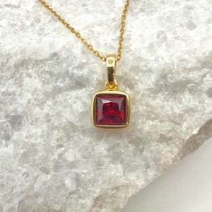 May include: A gold-colored necklace with a square, red gemstone pendant. The pendant is set in a gold frame and hangs from a delicate gold chain. The gemstone has a deep red color.