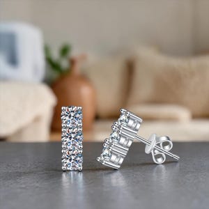 May include: A pair of silver stud earrings with three clear, sparkling stones set in a row. The earrings are displayed on a gray surface, with a blurred background of neutral tones.