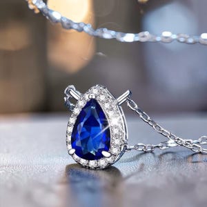 Puede incluir: Un collar de tono plateado con un colgante de piedra preciosa azul profundo en forma de pera. La piedra central azul está rodeada por un halo de pequeñas piedras claras y brillantes engastadas en un marco metálico, unido a una cadena fina.