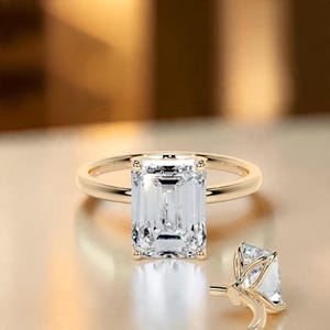 May include: A gold ring featuring a large, clear, emerald-cut center stone held by four prongs. The band is smooth and polished gold. A second, partial view of a similar ring shows the side profile of the stone setting and prongs.