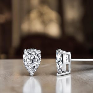 May include: A pair of pear-shaped diamond stud earrings. The diamonds are clear and faceted, set in a silver metal. The earrings are displayed on a light-coloured surface with a blurred background.