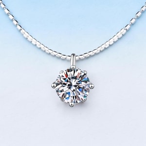 May include: A silver necklace with a round, clear gemstone pendant. The pendant is the focal point, reflecting light with multiple facets. The chain is delicate and silver, set against a gradient blue background.