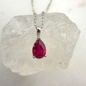 Op de afbeelding: Een zilveren hangerketting met een druppelvormige, helderrode edelsteen. De edelsteen is gezet in een zilveren bezel en hangt aan een fijne zilveren ketting. De ketting wordt getoond tegen een witte, getextureerde achtergrond.