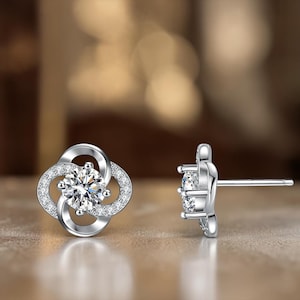 May include: A pair of silver-colored stud earrings. Each earring features a four-leaf clover design with a central, round, clear gemstone surrounded by smaller, clear gemstones. The earrings are set against a blurred, warm-toned background.
