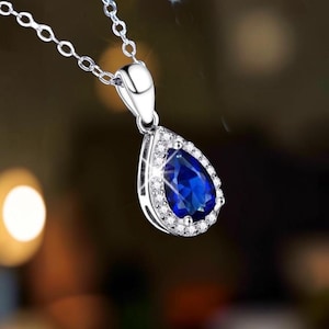 Puede incluir: Un collar con colgante en tono plateado que presenta una piedra preciosa azul intenso en forma de pera en el centro. La piedra está rodeada por un halo de pequeñas piedras claras y brillantes en un marco metálico, colgado de una cadena fina.