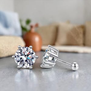 May include: Two silver stud earrings with large, clear, round-cut gemstones. Each earring features a six-prong setting and a post with a small ball backing. The earrings are displayed on a light gray surface.