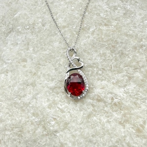 May include: A silver-toned necklace featuring a pendant with a large, oval-cut deep red gemstone at the centre. The pendant is accented with small, clear, sparkling stones along the frame and includes a heart-shaped design at the top bail.