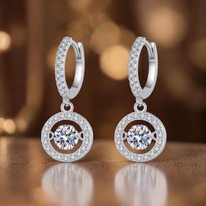 May include: A pair of silver hoop earrings with a round, sparkling, clear gemstone center. The gemstone is surrounded by a halo of smaller, clear stones. The earrings are set against a blurred, warm-toned background.
