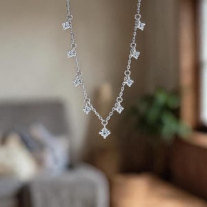 May include: A delicate silver necklace with multiple square-cut, clear gemstones. The necklace hangs gracefully, showcasing the sparkling stones against a blurred background. The design is simple yet elegant, suitable for various occasions.