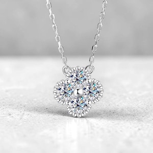 May include: A silver necklace featuring a four-leaf clover pendant. The pendant is adorned with sparkling, clear gemstones and a small, round center stone. The chain is delicate and the background is a soft, neutral color.