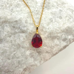 Può includere: Una collana color oro con un pendente a forma di goccia, sfaccettato, di colore rosso scuro. Il pendente è sospeso a una delicata catena. La collana è esposta su uno sfondo bianco strutturato.