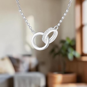May include: A silver-toned necklace featuring two interlocking circular pendants. One circle is a plain polished band, while the other is a slightly smaller band encrusted with small, clear, sparkling stones. The pendant hangs from a delicate chain.