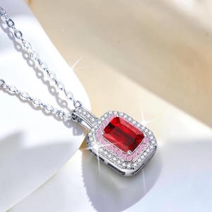 May include: A silver-toned necklace featuring a rectangular pendant with a large, deep red center stone. The stone is surrounded by a halo of small pink stones, followed by an outer border of clear white stones on a silver-toned metal setting.