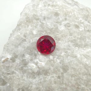 May include: A round, faceted, deep red gemstone. The gem is displayed on a white, textured surface. The cut of the gemstone creates multiple facets that reflect light, enhancing its brilliance. This image showcases a single, vibrant gemstone.