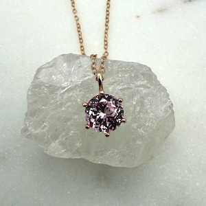 May include: A rose gold necklace with a round, pale pink gemstone pendant. The pendant is held by six prongs and hangs from a fine chain. The necklace is shown on a white, textured surface.