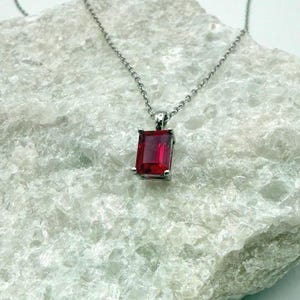 May include: A silver-toned necklace featuring a rectangular, emerald-cut red gemstone pendant held by four prongs. The pendant is suspended from a delicate, thin silver-toned chain that connects to the top of the metal setting.