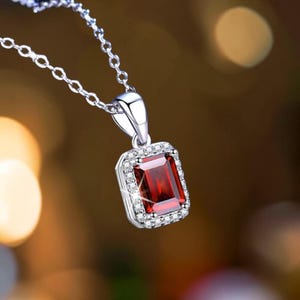 May include: A silver-toned necklace featuring a rectangular, emerald-cut deep red gemstone pendant. The central red stone is surrounded by a halo of small, clear, sparkling stones set in a metallic frame, suspended from a delicate silver chain.