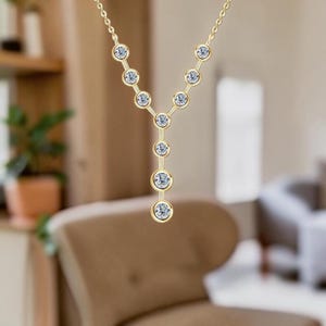 May include: A gold-tone Y-shaped necklace featuring a delicate chain and a series of bezel-set clear round stones arranged in a vertical drop pattern. The design consists of two diagonal lines of stones meeting at a central point, extending downward.