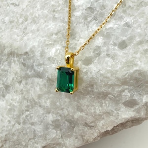 May include: A gold-colored necklace with a rectangular, emerald-green gemstone pendant. The pendant is set in a gold frame and hangs from a delicate gold chain. The necklace is displayed on a white, textured surface.